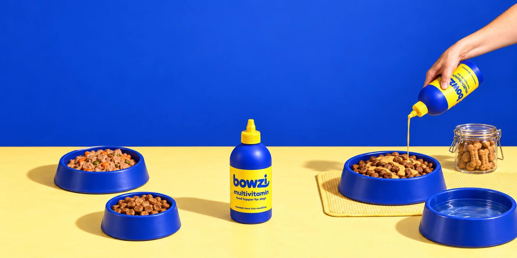 Bowzi on a yellow table with bowls of dog food and a hand pouring topper